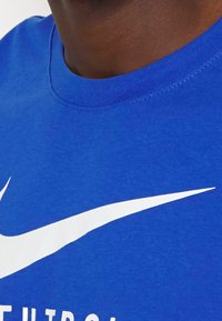 Zblízka na osobu v modré košili s bílým logem Nike swoosh a částečným textem na hrudi.