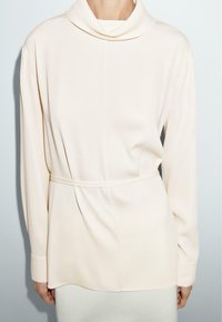 Haut à manches longues avec col drapé en tissu beige doux, doté d'une taille ceinturée et d'un drapé subtil, avec une texture lisse et une coupe décontractée.