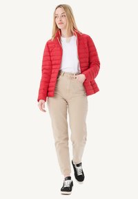 Giacca trapuntata rossa con colletto alto, indossata sopra una camicia bianca. Pantaloni beige a vita alta e sneakers nere con lacci bianchi.
