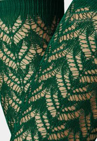 Grüne Spitze Leggings mit einem geometrischen Blattmuster, das komplexe Details und ein strukturiertes Design auf einem hautfarbenen Hintergrund zeigt.