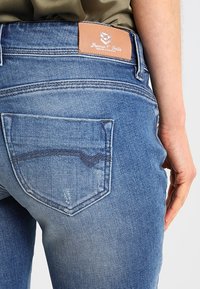 Blå denimjeans med ljus tvätt, subtil blekning och en läderlogotyp. Två bakfickor med kontrasterande stygn.