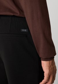 Primer plano de una persona que lleva pantalones negros con una pequeña etiqueta "Nineteen Ninety Three" cerca del bolsillo trasero y una camisa de manga larga marrón.
