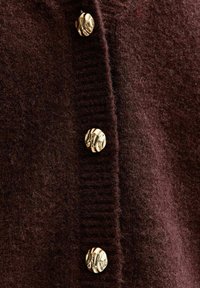 Burgunderröd stickad cardigan med texturerad, tjockt tyg och stora guldknappar längs framfickan. Ribbad nederkant och ärmslut.