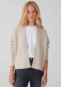 Cardigan beige chiaro realizzato in tessuto morbido e testurizzato, con apertura frontale, spalle scese e una vestibilità rilassata. Indossato sopra una camicia bianca.