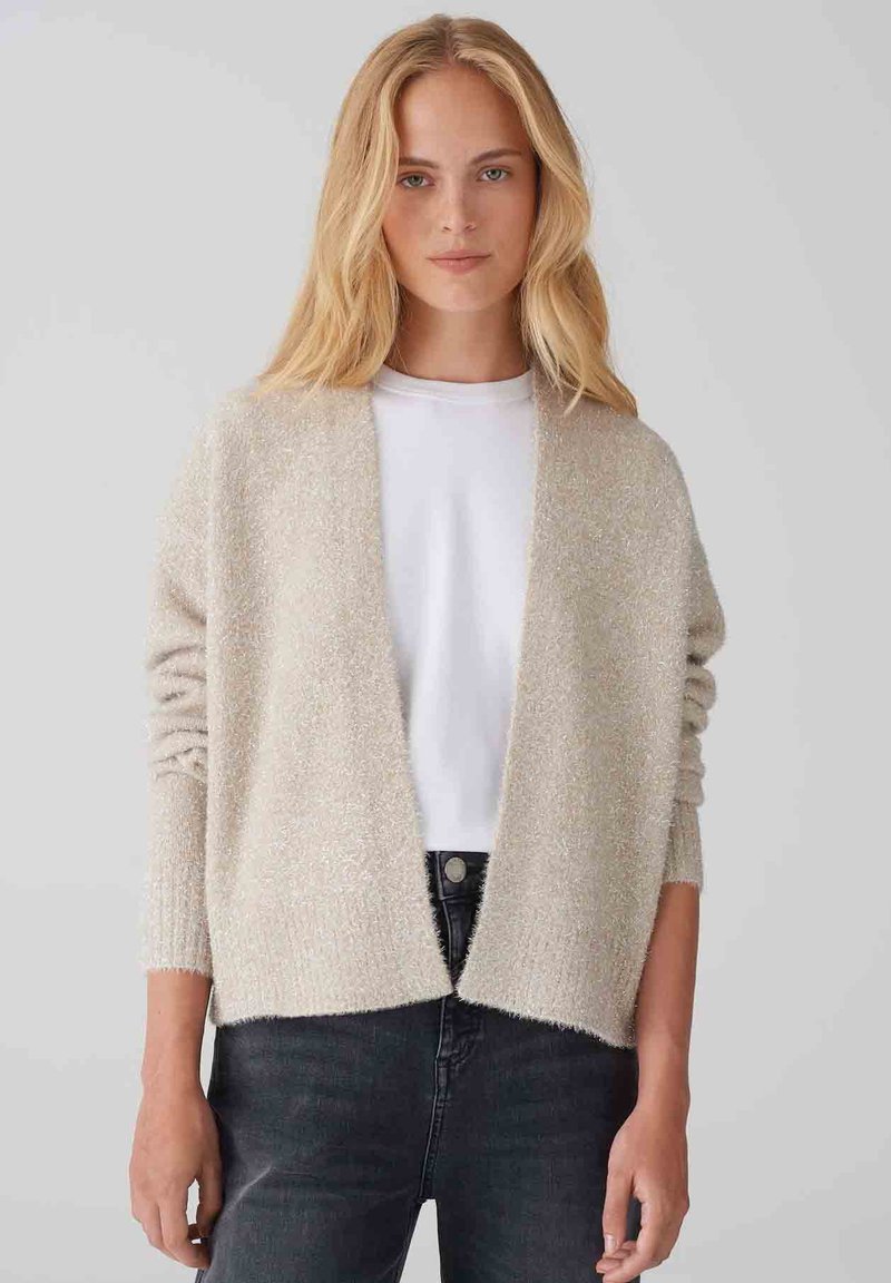 Cardigan beige chiaro realizzato in tessuto morbido e testurizzato, con apertura frontale, spalle scese e una vestibilità rilassata. Indossato sopra una camicia bianca.