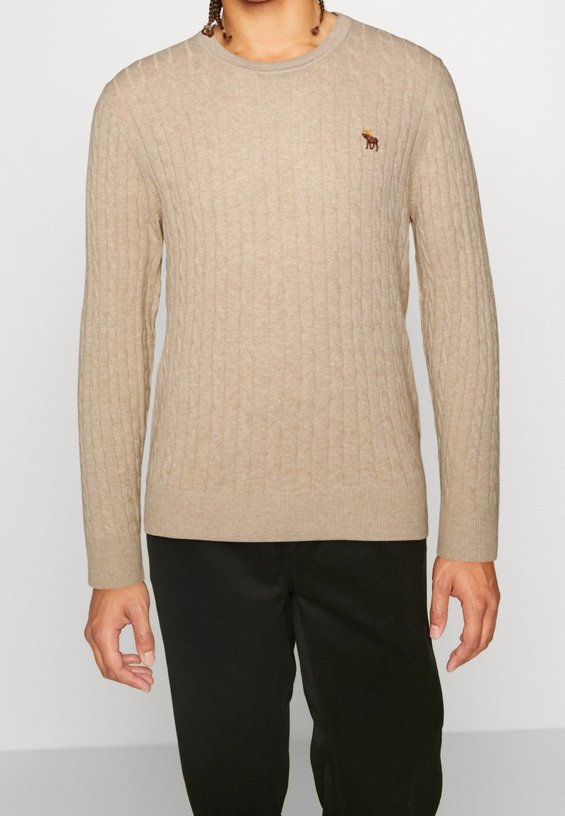 Mann trägt einen beigefarbenen, grobgestrickten Pullover mit langen Ärmeln und einem kleinen, gestickten Elch auf der Brust, kombiniert mit einer schwarzen Hose.