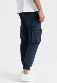Pantalons de cargo navy avec des chevilles élastiques, plusieurs poches zippées et une texture lisse, assortis à des baskets blanches.