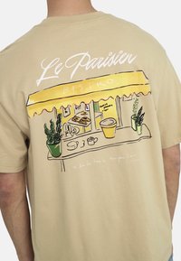 Beige Baumwoll-T-Shirt mit einer grafischen Illustration eines Bistros mit gelbem Sonnenschirm, Pflanzen, Tassen und Gebäck im Linienstil.
