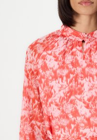 Bluse med blomsterprint i korall og pink, med høy krage, knappelukking og lange ermer med innsamlet detaljer.