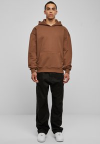 Brun oversized hettegenser med kengurulomme, ribbede mansjetter og snor i hetta, kombinert med svarte bukser og hvite joggesko.
