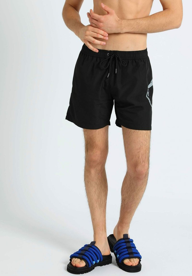 Pantaloni da bagno neri con vita elasticizzata e cordoncino, caratterizzati da un design geometrico verde sul lato. Indossati con sandali blu con cinturini.