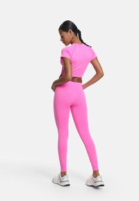 Prilagođeni ružičasti sportski top i tajice od glatkog, rastezljivog materijala. Model prikazan s leđa, u kombinaciji s bijelim sportskim cipelama.