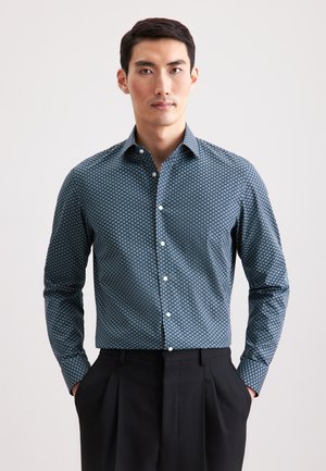 Blaues Hemd mit geometrischem Muster, langen Ärmeln und spitzem Kragen, kombiniert mit schwarzen, plissierten Hosen.