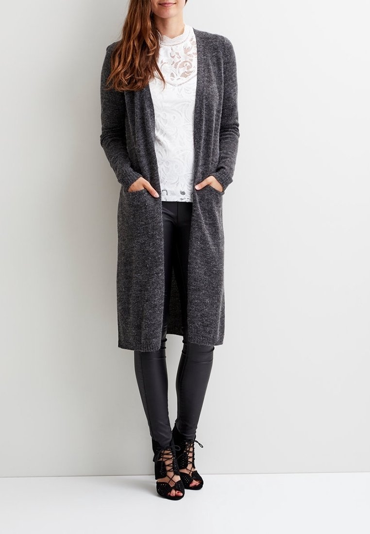 Lång grå cardigan med öppen front, ribbad textur och sidofickor. Bärs över en vit topp och svarta tajta byxor, kompletterad med svarta snörda klackar.