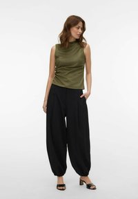 Femme portant un haut sans manches vert olive à col haut, un pantalon noir ample avec ourlet froncé, et des sandales noires à talons blocs ouverts, debout.