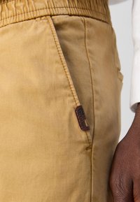 Pantalon en coton beige avec une taille élastique, une poche latérale et une étiquette en cuir marron avec le détail "Est. 1993". Texture lisse.