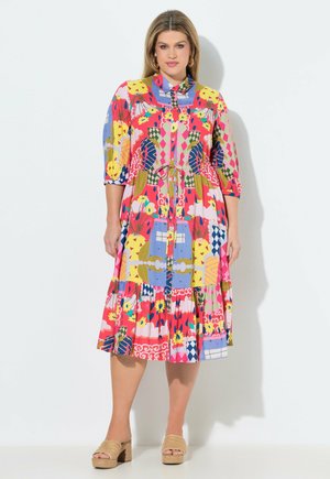 Donna che indossa un vestito midi colorato e a fantasia con maniche a tre quarti e sandali beige con zeppa, in piedi su uno sfondo bianco semplice.