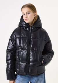 Zwarte pufferjas met een glanzende afwerking, geblokt ontwerp, oversized capuchon en een rits aan de voorkant. Draag het over een spijkerbroek.