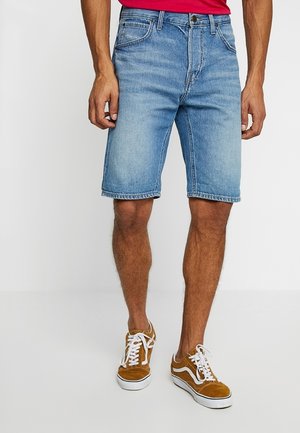 Man die lichtblauwe jeansshorts, een rood shirt en bruine sneakers met witte veters draagt, staand op een witte vloer.