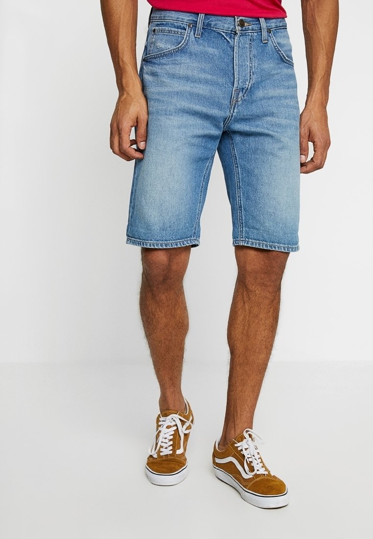 Homme portant un short en jean bleu clair, une chemise rouge et des baskets marron à lacets blancs, debout sur un sol blanc.