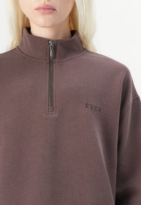 Brun zip-up genser laget av teksturert stoff, med høy krage og en liten logo på venstre side.