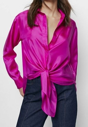 Vrouw draagt een felroze satijnen blouse met lange mouwen, gestrikt in de taille, gecombineerd met donkerblauwe spijkerbroek.