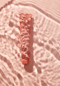 Vaaleanpunainen putki, jossa on etiketti "Indie Lee Cleansing Balm", osittain veden pinnassa, pehmeällä ja kiiltävällä pinnalla, jonka pinnalla on lempeitä aaltoja heijastamassa valoa.