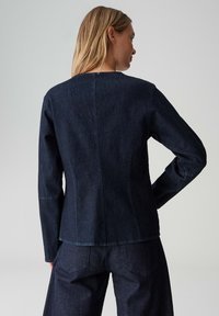 Dunkle Jeansjacke mit langen Ärmeln. Verfügt über einen geraden Schnitt und schlichte Nahtdetails. Glatte Textur und abgerundeter Ausschnitt.