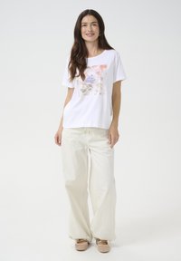 T-shirt blanche avec un design graphique floral en rose et bleu, associée à un pantalon en crème ample et des chaussures à enfiler beiges.