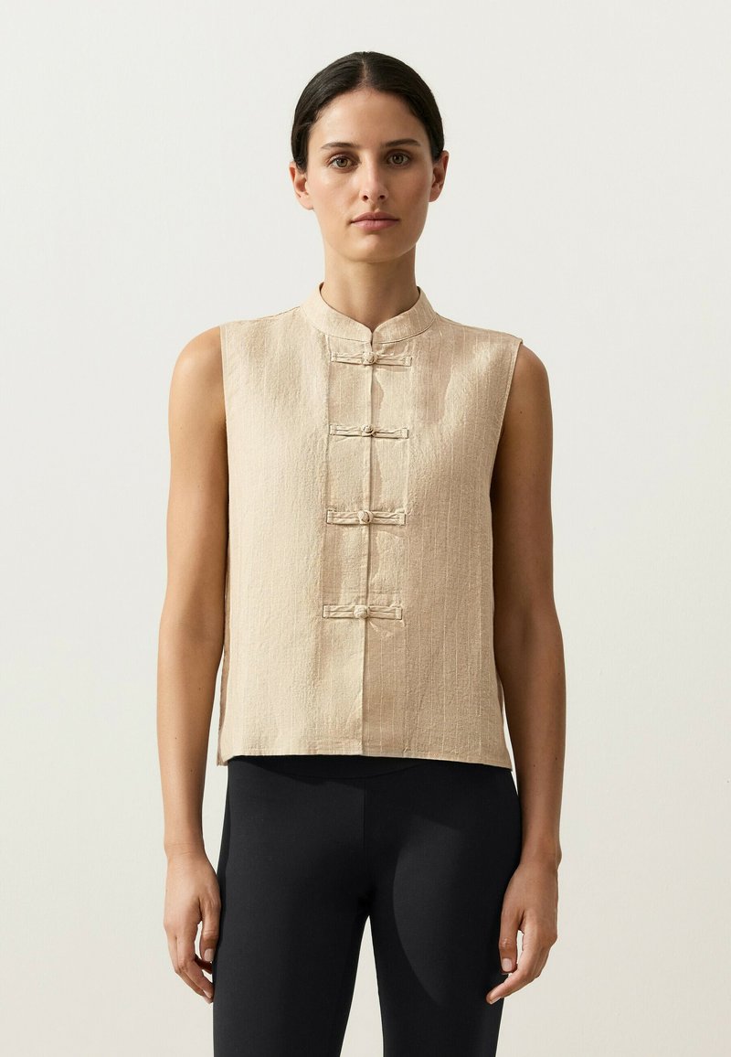 Femme portant un haut beige sans manches à col mandarin avec des fermetures à nœuds chinois, associé à un pantalon noir moulant, debout devant un fond uni.