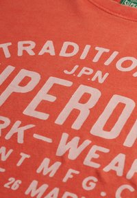 Nærbillede af rødt stof med hvidt trykt tekst, inklusive "TRADITION," "J.P.N," "SUPER," og delvise ord relateret til arbejdstøj.
