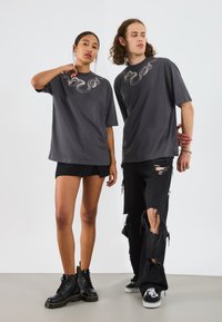 T-shirts gris surdimensionnés avec un design de serpent, associés à un jean noir déchiré et des bottes noires. Les shorts du modèle sont noirs ; ils ont un look superposé.
