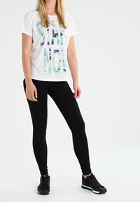 T-shirt blanc avec "START NOW" imprimé en lettres multicolores, associé à un legging noir et des chaussures de sport noires. Design simple, coupe décontractée.