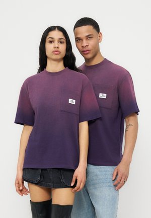 Dwa modele noszące dopasowane, fioletowe koszulki typu oversize z gradientem i kieszeniami na piersi, jedna w połączeniu z czarną spódnicą i botkami, druga z jasnymi dżinsami.