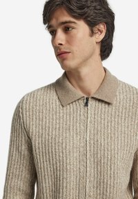 Beige ribbad stickad tröja med dragkedja fram och en mjuk, kontrasterande krage. Texturen är mysig och inbjudande, med en strukturerad silhuett.