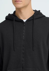 Sweat à capuche noir zippé en tissu doux, avec capuche à cordon, fermeture éclair et poches avant, présentant une texture lisse et un design minimaliste.