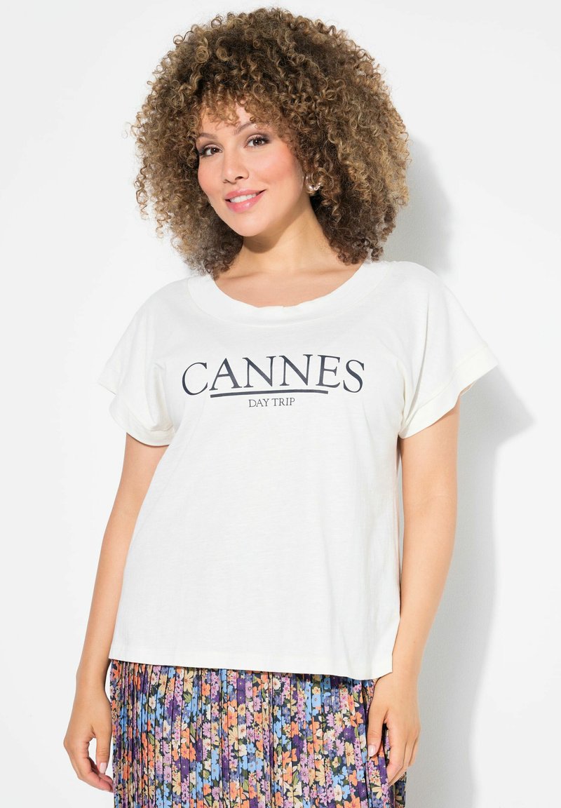 Camiseta blanca de manga corta con el texto "CANNES DAY TRIP" en azul marino, combinada con una falda plisada floral multicolor. Textura de tela suave.