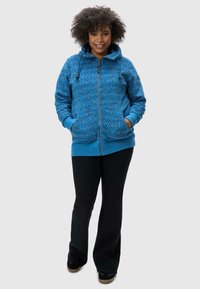 Haut en tricot bleu avec une fermeture éclair à l'avant, featuring des cordons de serrage doubles, un motif texturé et des poignets côtelés. Associé à un pantalon évasé noir et des chaussures.