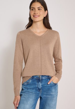 Kvinde iført en beige langærmet V-hals trøje og blå jeans, smilende let med den ene hånd i lommen mod en neutral baggrund.