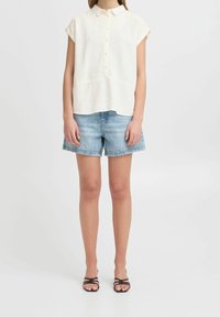 Donna che indossa una camicia in crema a maniche corte con bottoni, pantaloni corti in denim azzurro chiaro e sandali con tacco neri a cinghietti, in piedi su uno sfondo bianco.