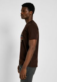 Camiseta marrón de manga corta con cuello redondo, que presenta un estampado gráfico en colores brillantes. El tejido tiene una textura suave. El modelo lleva puestos unos jeans grises.