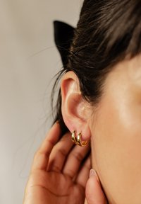 Boucles d'oreilles en or avec un design courbé et une finition polie, portées sur un lobe d'oreille. Les cheveux sont tirés en arrière, mettant en valeur les boucles d'oreilles.