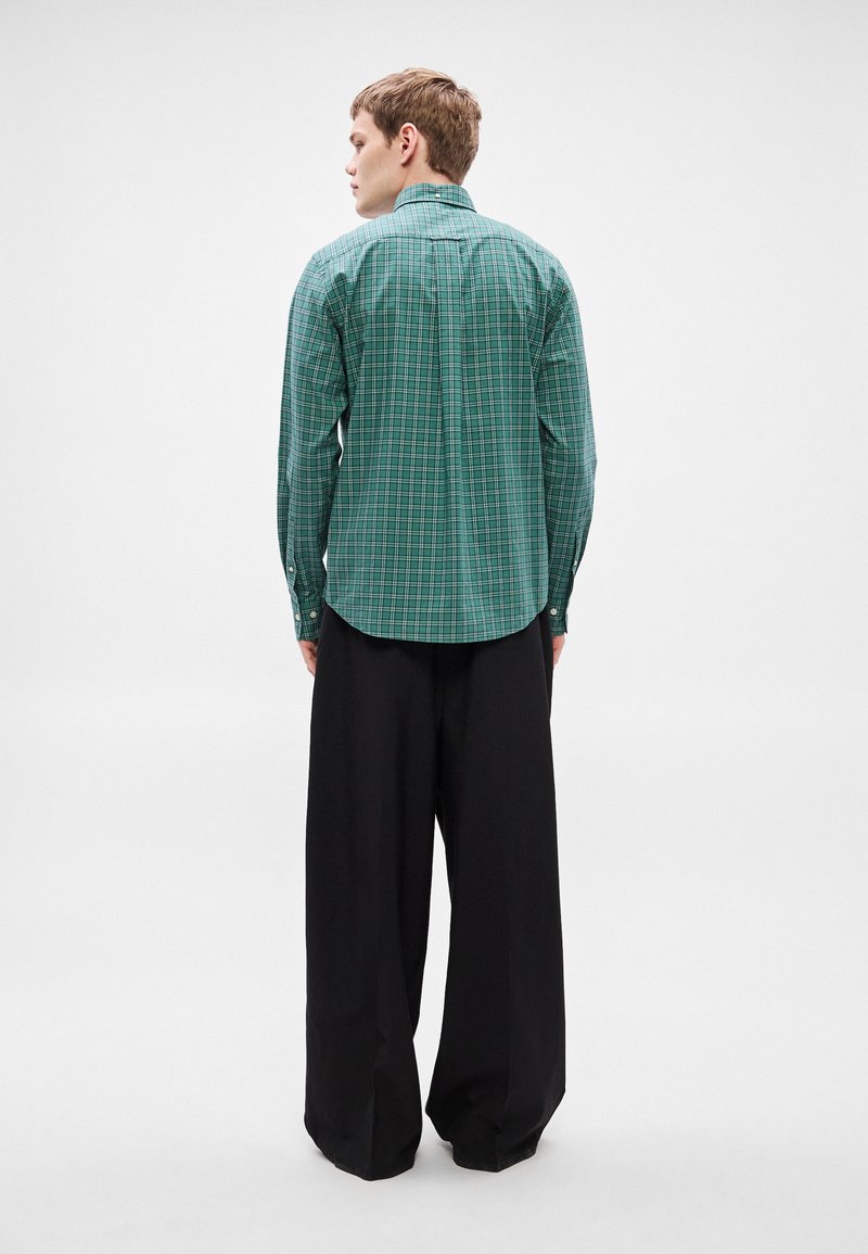 Green plaid shirt with a curved hem and button cuffs, paired with loose black trousers. The fabric appears lightweight and textured.