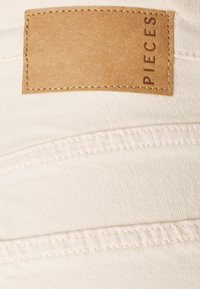 Krämfärgad tyg med sydda detaljer och en rektangulär, tanfärgad läderlapp som visar "PIECES" präglad i brunt.