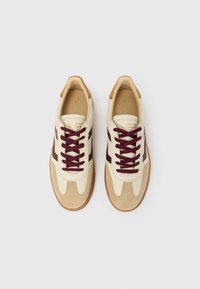 Paire de baskets beiges et marron avec des lacets bordeaux, des bouts en daim et le nom de la marque visible sur la languette et les côtés, sur fond blanc.