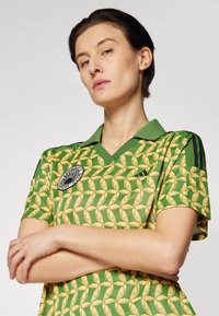 Žalias polo marškinėliai, pagaminti iš lengvos medžiagos, su geltonų bananų raštu, juodomis trimis juostelėmis akcentais ir apvaliu logotipu ant krūtinės.
