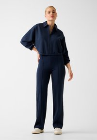 Korte marineblauwe jas met rits, gecombineerd met nauwsluitende marineblauwe broek. Beide gemaakt van gladde stof, met strakke lijnen en minimale details.