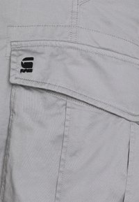 Light gray fabric features a prominent black logo. The garment has a pocket with a flap and detailed stitching, showcasing a smooth texture.