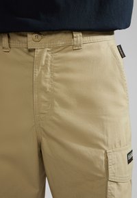 Pantalones cargo beige con una textura suave, que cuentan con un cierre frontal de botón, bolsillos laterales y una etiqueta de marca en el bolsillo.