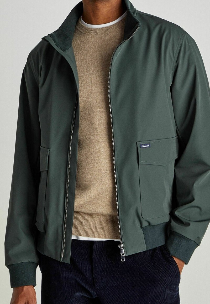 Bomber jacket verde con chiusura zip, polsini a costine e due tasche frontali. Indossato sopra un maglione beige. Texture liscia e opaca.